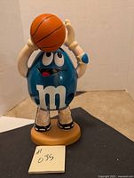 Front view of the blue M&M dispenser figurine holding a basketball above head, showing details of the face, gloves, and shoes.