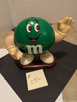 Full view of green M&M candy dispenser standing on brown base, waving hand, smiling face with big eyes
