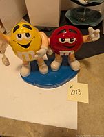Full view of the red and yellow M&M characters standing on a blue base, showing their facial expressions and posed hands.