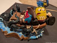 Yellow M&M pirate figurine seated on a small brown boat with black tentacles and blue wave base.
