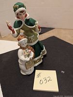 Porcelain doll dressed in green and white Victorian-style clothing holding two small bottles, with a smaller white figure holding a gift bag integrated in front.