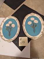 Two oval wall plaques with sculptured white flowers on blue textured backgrounds, cream ornate frames. Both plaques shown side by side.