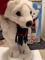 Close-up image of the white plush polar bear figurine wearing the striped scarf with black, red, and green colors, showing detailed face and front paws, with the product box visible behind.