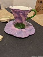Front angle photo of purple flower teacup on matching saucer, showing green handle and petal details.