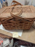 Front angled view of antique wicker picnic basket showing the woven basket body, hinged wooden slatted lid, and crossed wooden handles resting on top.