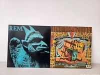 Two R.E.M. vinyl record album covers, Chronic Town with blue gargoyle and Reconstruction with theatrical stage artwork.