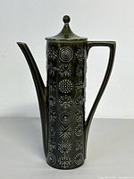 Front left angled view of tall ceramic coffee pot with embossed Totem pattern designs and dark green glaze.