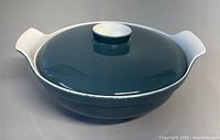 Side angle view of teal blue lidded ceramic bowl with white handles and white lid knob, showing shape and color of bowl.
