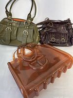Three handbags displayed together on a white background showing front view including the large translucent jelly shopper and two leather handbags.
