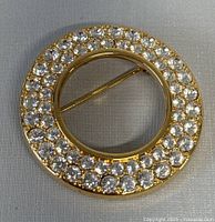 Top view of round gold-tone brooch with two rows of clear Swarovski crystals against a white background, showing pin closure on back.