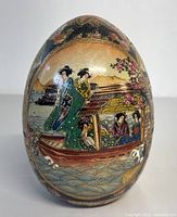 Front view of hand painted Satsuma egg showing geishas near a boat and water with floral and mountain background.