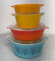 Stacked view of four vintage casserole bowls in yellow, orange, and blue with clear lids on two.