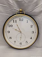 Front view of the large wall clock showing white face, black Arabic numerals, gold rim, and clock hands. Smaller inner dial also visible.