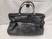 Front view of the black travel bag showing structured shape, geometric pattern on fabric, and leather handles wrapped in plastic.