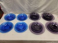 Blue and purple glass dishes arranged in two groups on a white cloth background. Four blue dishes (2 bowls, 2 plates) on left and four purple dishes (2 bowls, 2 plates) on right.