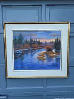 Framed landscape painting showing a river flowing through a forest with sunset sky, housed in a gold-tone frame with white matting, photographed against a blue garage door.