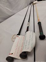 Four horse whips arranged side by side on a white surface, showing overall condition and attached tags with horse and jockey information.