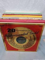 Photo showing a stack of vinyl records with the top record titled '20 Instrumental Hits Instrumental Gold' with a red and gold album cover featuring a vinyl record label design.