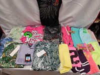 Overview of the lot of young ladies new clothing items with tags, showing 13 clothing pieces including tops, shorts, and pants in various colors and prints.