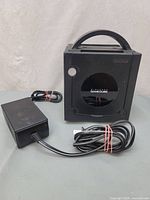 Nintendo GameCube console with power adapter visible on table, console facing forward showing logo and power button.