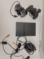 Two wired PlayStation 2 controllers and PS2 slim console with power and AV cables laid out on white background.