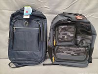 Two backpacks standing upright side by side on a bed or soft surface. Left backpack is dark blue with tags attached, right backpack is black and grey camo patterned with multiple pockets.