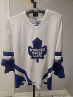 White Toronto Maple Leafs CCM hockey jersey hanging on a hanger, showing front with large blue Maple Leaf logo and blue and white sleeve stripes.