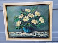 Oil painting showing full view of bouquet of white chrysanthemum-like flowers in a blue vase on a white cloth surface, framed in light wood.