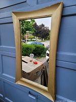Mirror hung on an outdoor garage wall showing the gold frame and reflection of surrounding outdoor scene including a driveway with cars.