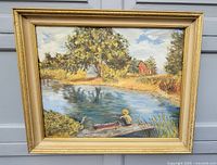 Front view of framed painting depicting person fishing by a dock on a river with rural landscape including trees and a red house in background.