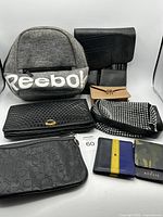 Photo showing the full assortment of bags and wallets on a white background including the Reebok backpack at rear left, black quilted clutch, black studded handbag, Calvin Klein clutch, small tan wallet, and two small wallets in front row.