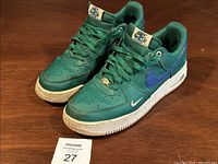 Pair of green Nike Air Force One sneakers on wooden surface, showing overall worn condition and dirt on white soles.