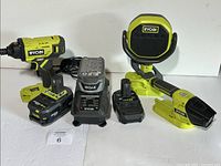 Full view of Ryobi impact driver with battery attached, Bluetooth work light speaker, battery charger, and three batteries arranged on table.