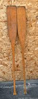 Two wooden rowing oars upright against plywood background showing full length and handles.