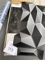 Top-down view of black faceted 3D wall panel showing geometric polygonal design with multiple facets creating a three-dimensional look. Panel is lying flat on a cardboard box with a paper label.