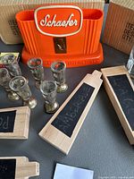 Photo of vintage Schaefer orange plastic bar caddy, six cordial glasses with metal bases, four wooden paddle chalkboards with black chalkboard panels and internal chalk writing.