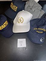 Photo showing four baseball caps with embroidered text 'Police Investigation', 'Probation', 'San Francisco Police', and a white cap with gold emblem, all on a table with striped cloth cap visible.