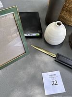 Photo of green enameled picture frame with gold accent, white pottery vase, black metal trinket box, black pen case with gold metal opener, and part of woven basket
