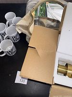 Six white mugs with black 'K' logo, IKEA mirror in gold frame, part of grey embroidered blanket, all placed in an open cardboard box.