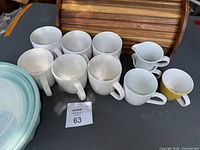Eight white ceramic mugs and one yellow ceramic mug arranged on a surface next to a wooden bread box
