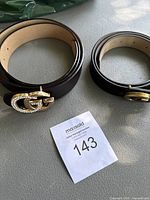 Photo showing two black leather belts with gold-tone double G buckles. One buckle is embellished with rhinestones, the other is plain gold-tone.