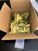 Open cardboard box containing multiple rubber frogs in green, yellow, and brown colors, some larger and some smaller, taken from top-down view.