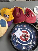Photo of three sports hats, a worn Olympus soccer ball, and a Chicago Cubs wall clock showing time 06:15. Hats include yellow Lakers, maroon Trojan, and faded maroon with '61'.