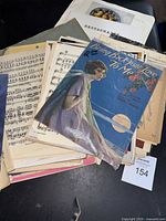 Top view of a pile of vintage sheet music and paper ephemera with a blue cover showing a woman and the title "Bring Back Your Love to Me." Items show wear and yellowing.