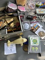 Overview of lot showing metal check file box open with assorted vintage items around it including signed golf balls in plastic cases, coins, costume goggles, pens, small figures, candy package, and jewelry.