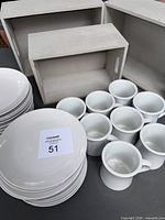Photo showing stacks of white IKEA plates in two sizes and ceramic mugs arranged in front inside grey wood storage boxes.