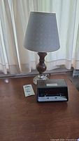 Table lamp with wood and hobnail glass base, white lampshade, and Sears alarm clock on wooden surface next to a manual or booklet.