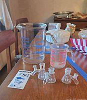 Photo showing 6-inch blue measured beaker, 3-inch red measured beaker with stirring rod, and four Warburg flasks with glass stoppers on wooden table.