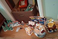 Photo showing several ceramic Christmas village houses with snow-covered roofs, small Christmas tree figurines, and boxed lighted Christmas village house.