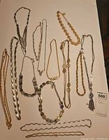 Overhead photo of thirteen assorted silver-tone and gold-tone costume necklaces arranged on a white surface showing variety in styles and lengths.
